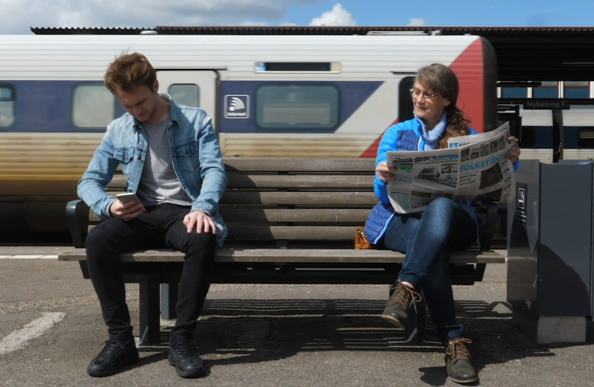 Ung mand med mobil sidder på en bænk på en togstation. Ved siden af sider en ældre kvinde med en Folketidende avis og smiler mens hun kigger mod hans telefon.